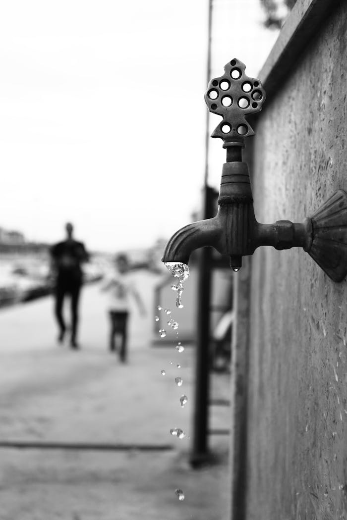 Black and white photo of a dripping faucet on a street with blurred people in the background.