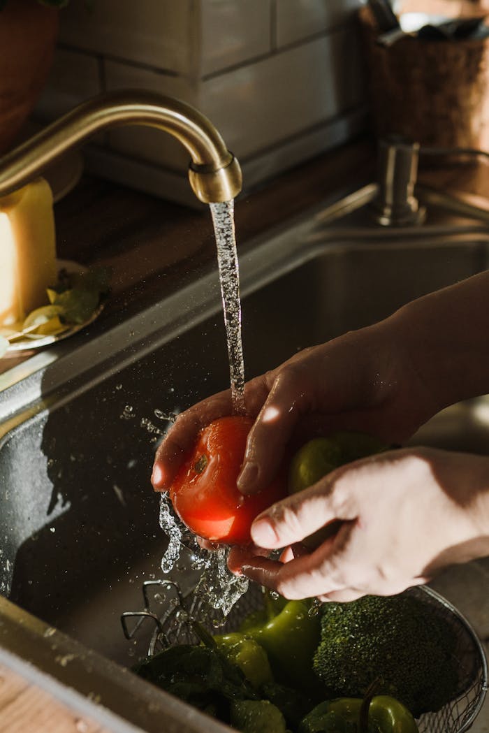 gallery-5 Close-up of hands washing a ripe tomato under a kitchen faucet. Perfect for themes of cleanliness and healthy living.